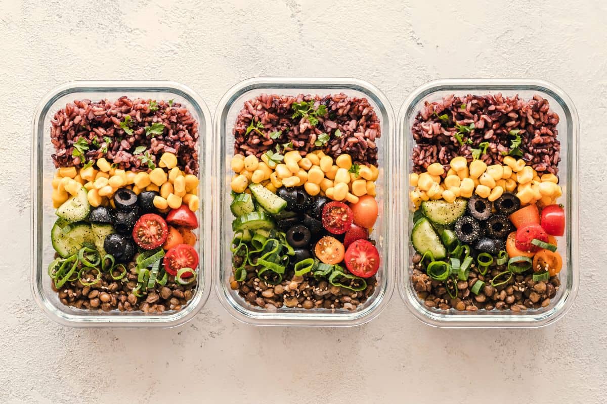 Three meal prep containers with rice, lentils, corn, olives, and vegetables