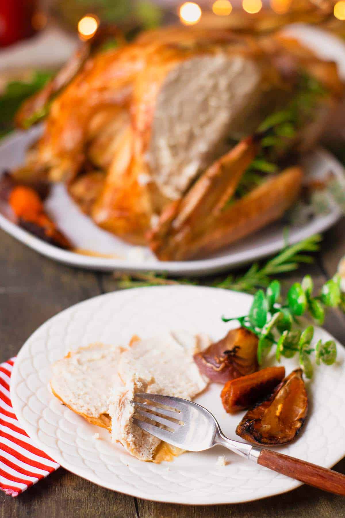 Sliced roast chicken on plate with fork and roasted vegetables, whole chicken behind.