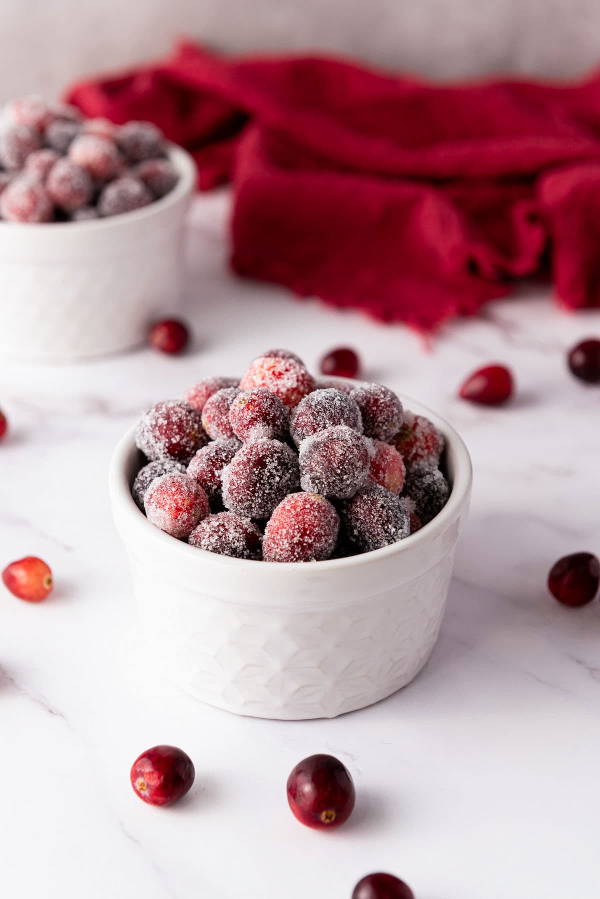 White bowl filled with sparkling sugared cranberries on a marble surface with red cloth.