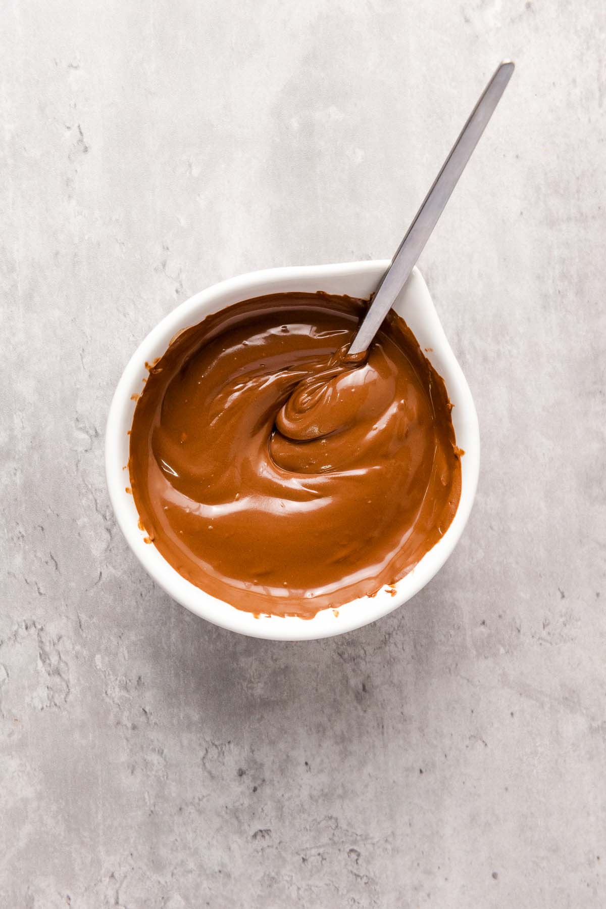 Bowl of melted chocolate with a spoon on a gray surface.
