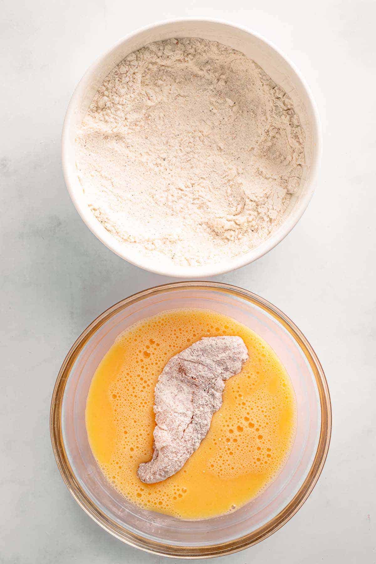 Chicken tender dipped in beaten egg beside a bowl of seasoned flour