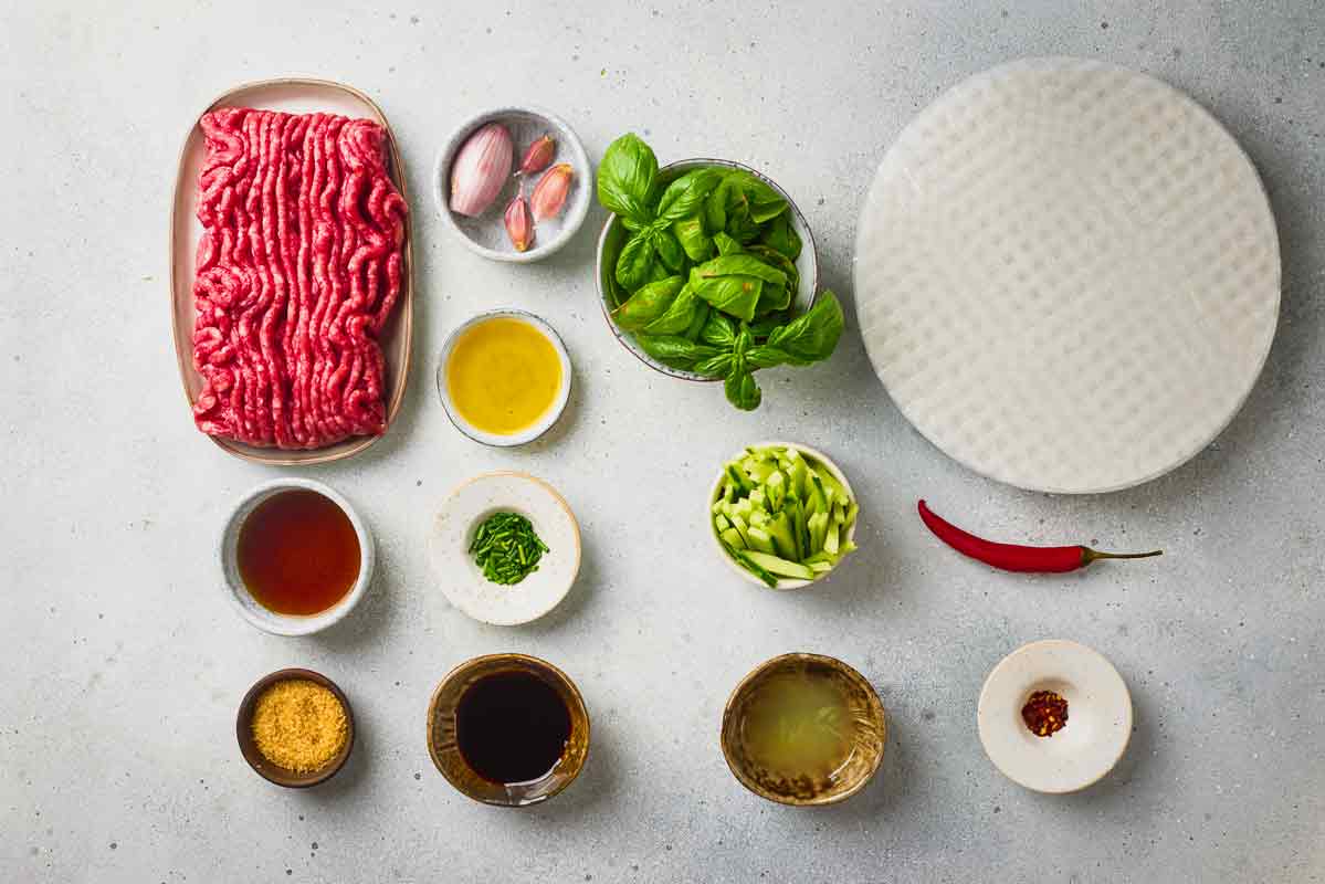 Ingredients for Thai-style ground beef spring rolls with rice paper, beef, basil, cucumber, and seasonings.