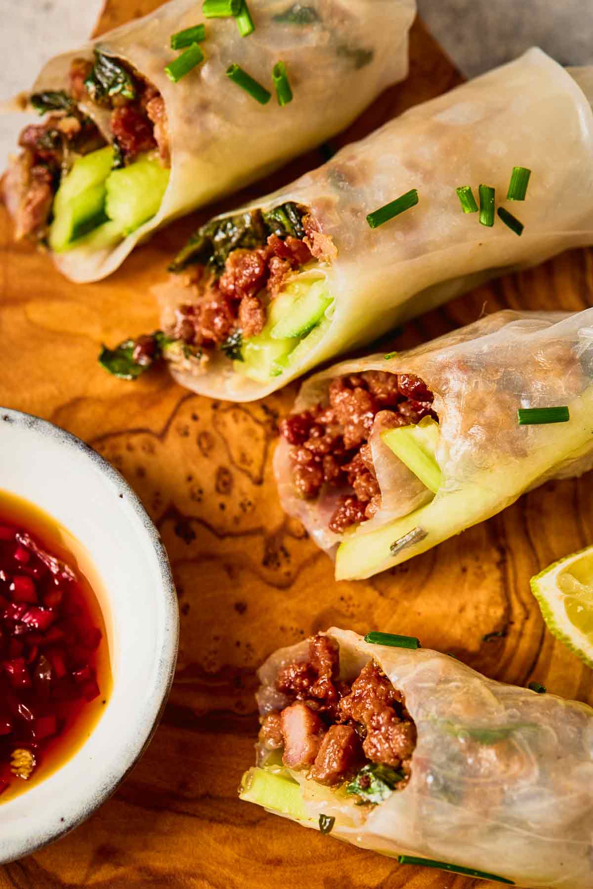 Thai-style ground beef spring rolls with cucumber, basil, and dipping sauce on a wooden board.