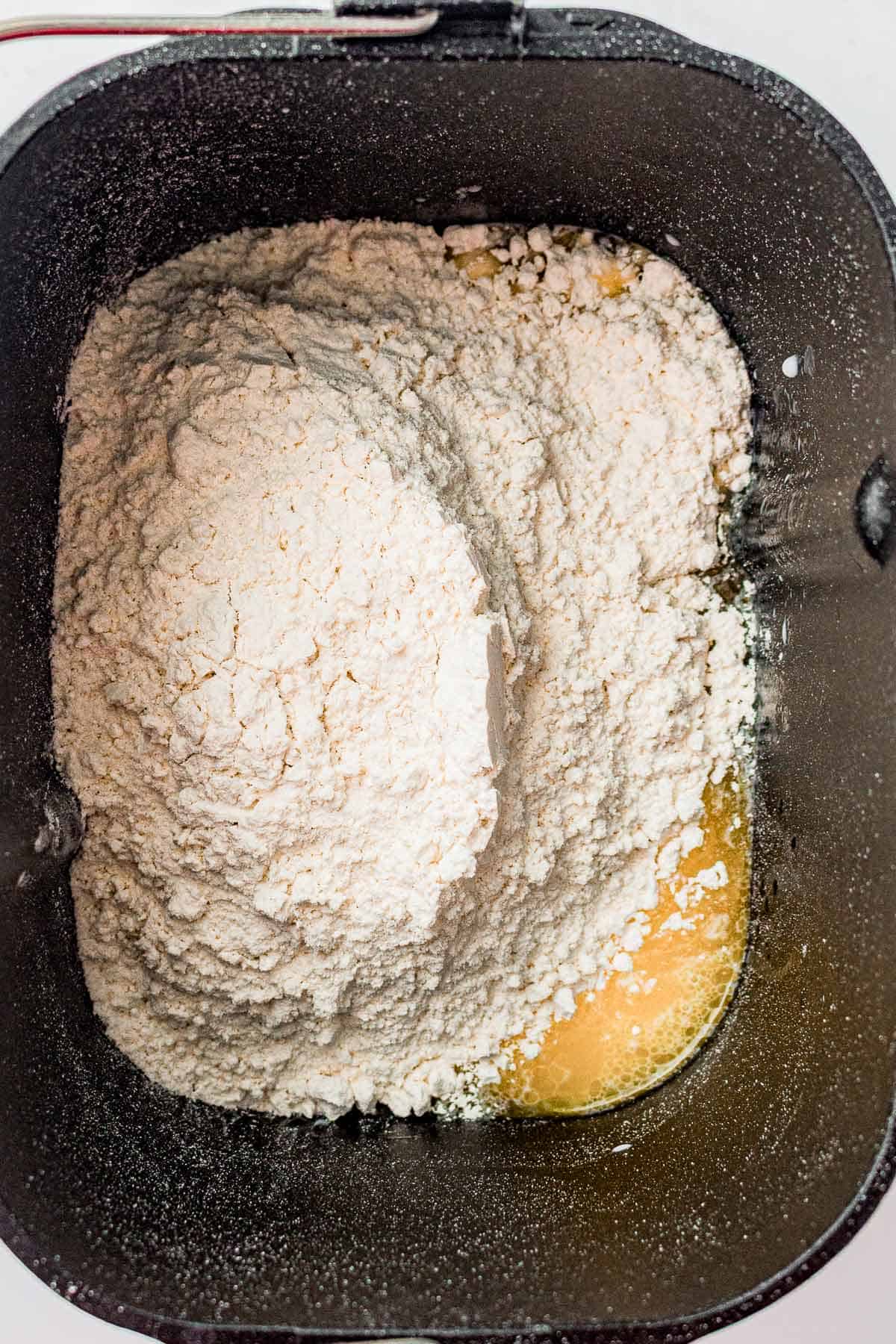 Bread machine pan with flour covering the wet ingredients before mixing.