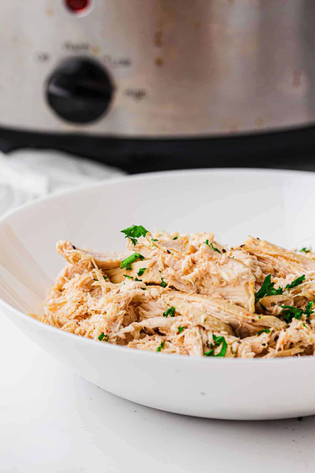 Bowl of slow cooker pulled chicken with a crockpot in the background.